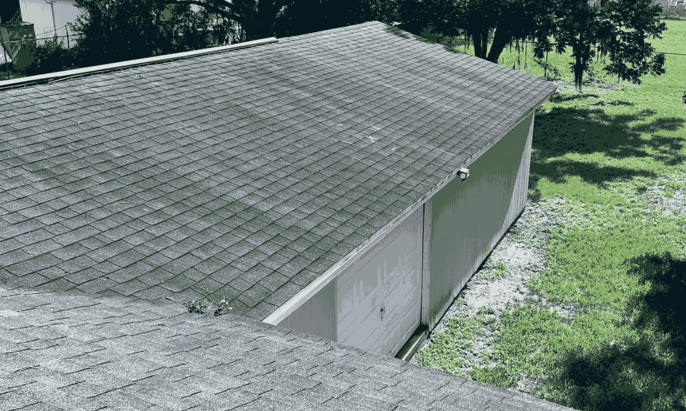 A gray shingled roof of a detached garage, expertly installed by Aleman Roofing, is pictured next to a patchy green lawn, viewed from above.