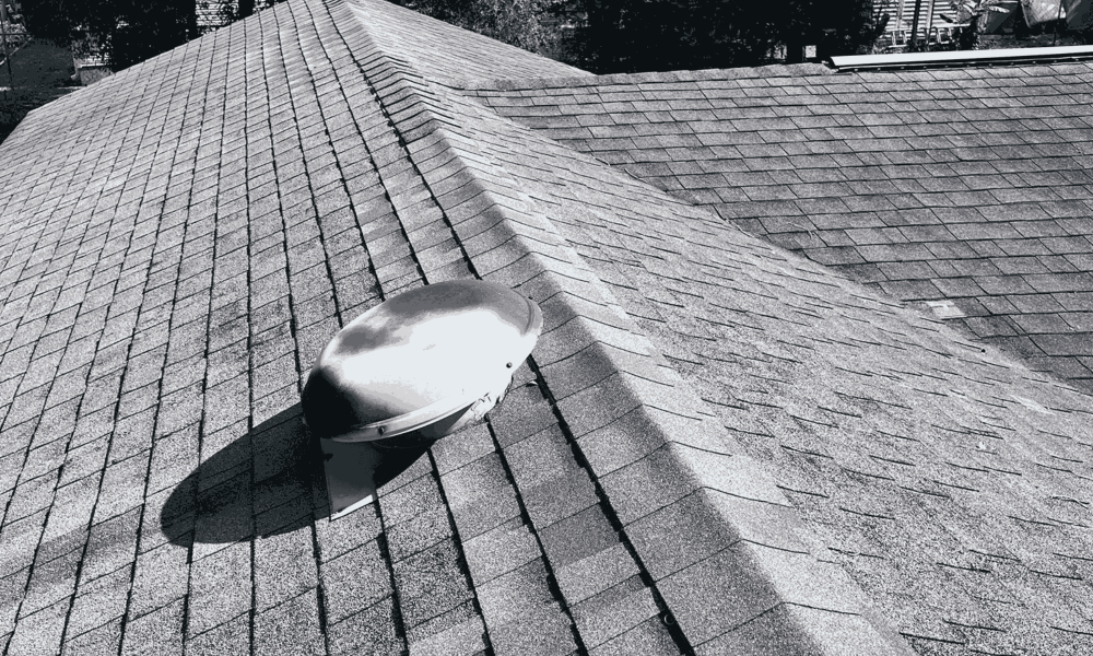 A rooftop with asphalt shingles, expertly installed by Aleman Roofing, features a round metal attic vent casting a shadow; trees and part of another roof are visible in the background.