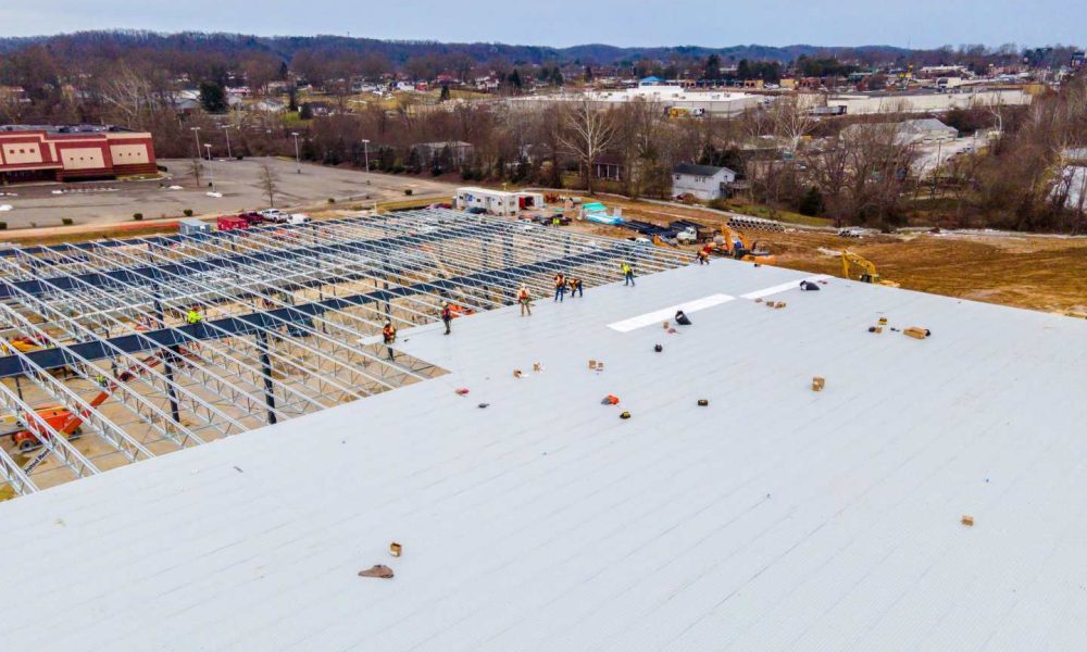 Aerial view of construction workers assembling a large metal roof structure at a commercial building site, with surrounding roads and buildings visible in the background.