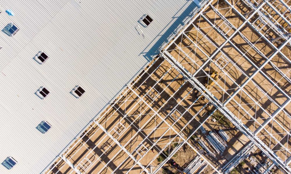 Aerial view of a construction site showing a partially built steel framework next to a completed section with a corrugated metal roof and several rectangular openings.
