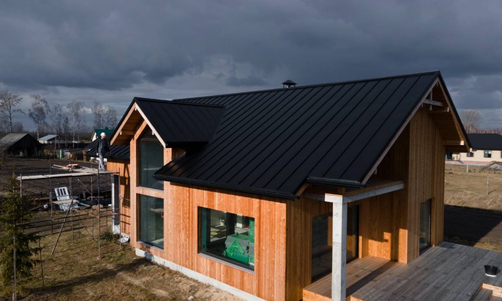 A modern wooden house with a black metal roof by Aleman Roofing stands on a grassy plot under a cloudy sky, featuring a large window and an unfinished deck.