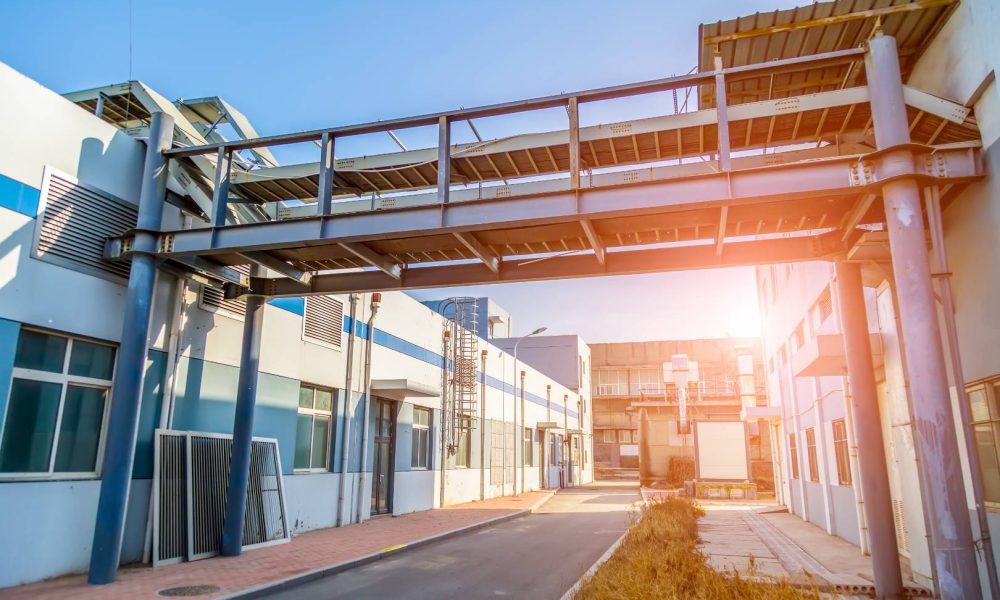 A modern industrial facility, about Aleman Roofing, with connected buildings and a skywalk above an empty paved pathway, under clear blue skies.