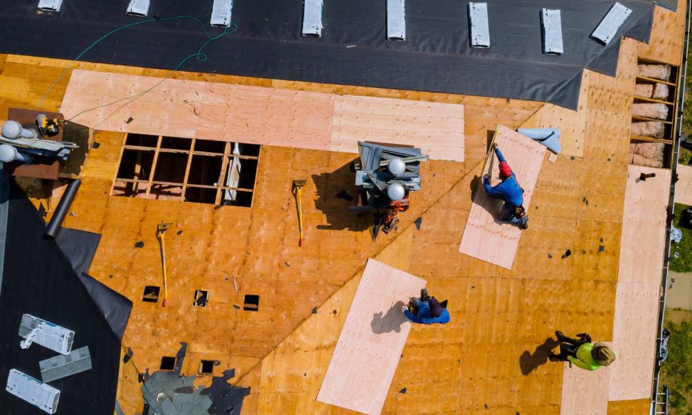 Aerial view of several workers installing plywood sheets and roofing materials on the roof of a building under construction.