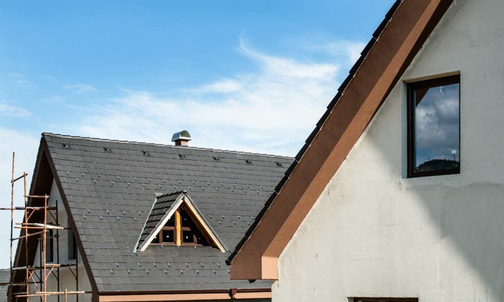 Two modern houses with dark shingle roofs and cream-colored walls under a blue sky; one house, featuring scaffolding on its side, highlights the quality craftsmanship about Aleman Roofing.