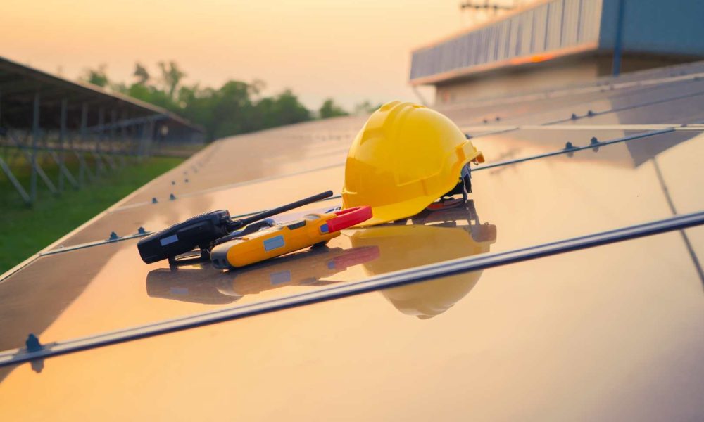 Yellow hard hat, safety glasses, and tools rest on solar panels, with a field and building visible in the background during sunset.