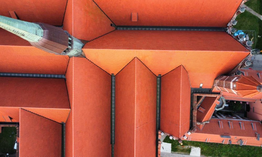 Aerial view of a large building with multiple pitched red tile roofs and a prominent spire, surrounded by green grass and walkways.