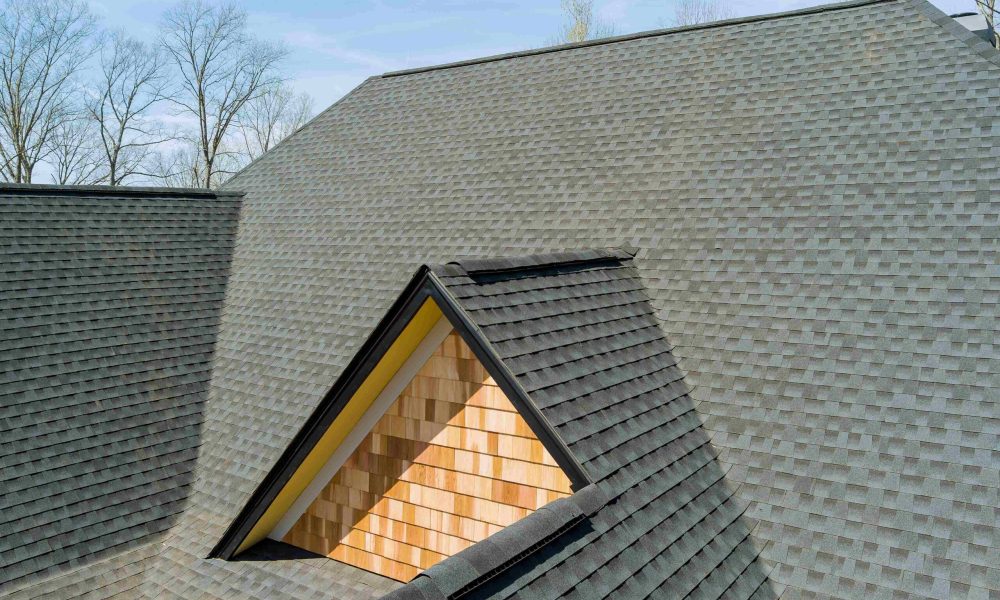 A close-up view of a house roof with gray asphalt shingles and a triangular dormer featuring wooden siding, expertly installed by Aleman Roofing, your trusted Central Florida Roofing Contractor. Leafless trees stand against a blue sky in the background.